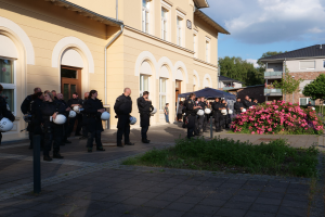 Eine Gruppe von Polizeibeamten steht vor einem Gebäude mit Fenstern und Türen, hält Helme in den Händen, mit einem Zelt in der Nähe, umgeben von Pflanzen mit Blumen, Gras, Bäumen und einem Straßenpfahl unter einem bewölkten Himmel.