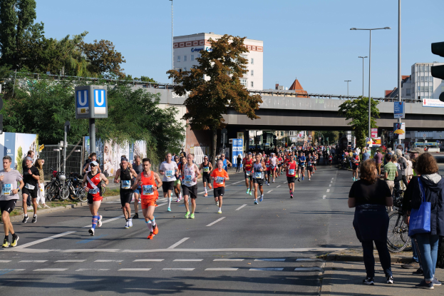 Gruppe von Menschen, die bei einem Marathon auf einer Straße mit Bäumen, Schildern und Laternen unter einem klaren blauen Himmel laufen.