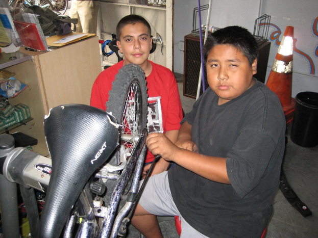 Zwei Personen reparieren ein Fahrrad an einem unordentlichen Schreibtisch mit Akten, Taschen und Kartons; Fahrräder, Ersatzteile, ein Schrank und andere Gegenstände sind im Hintergrund sichtbar.