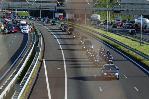 Eine belebte Autobahn mit Autos und Motorrädern, gesäumt von Geländern und Gras, mit Bäumen, Pfählen, Schildern und einer Brücke im Hintergrund.