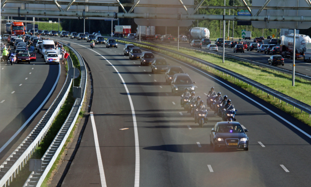 Eine belebte Autobahn mit Autos und Motorrädern, gesäumt von Geländern und Gras, mit Bäumen, Pfählen, Schildern und einer Brücke im Hintergrund.
