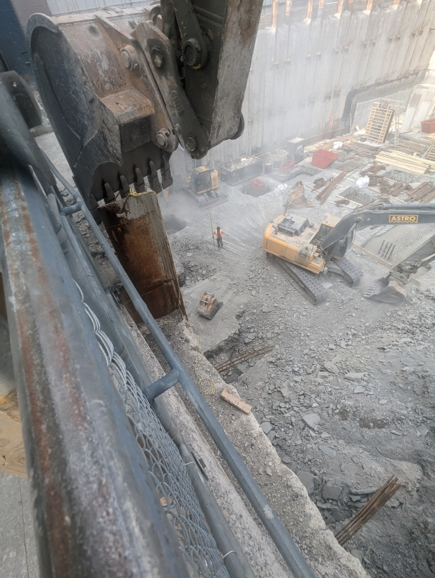 Eine Baustelle mit einem Bagger, der ein Gebäude abbricht, umgeben von Fahrzeugen, Menschen und einem Metallobjekt links.