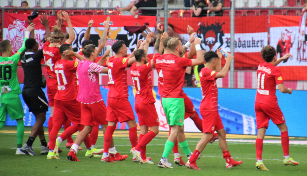 Eine Gruppe von Füßballspielern, die ein Tor feiern, einige halten Flaschen und andere klatschen, mit einem Zaun, Bannern, Metallstäben, Stühlen und einer Decke mit Deckenlampen im Hintergrund.