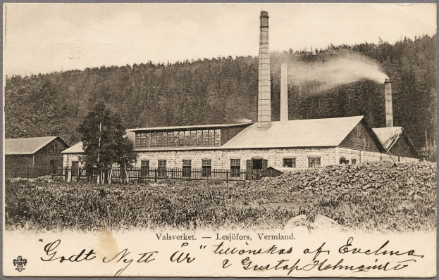 Schwarz-weiß-Fotografie einer Fabrik mit Rauchfahnen, umgeben von Häusern, Grünflächen und einem Zaun, mit Text unten.