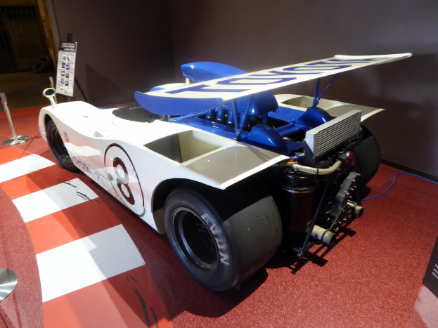 Ein weiß-blaues Rennfahrzeug in einem Museums Setting umgeben von Barrieren und Informationsschildern, mit einer Wand im Hintergrund, die auf das Goodwood Festival of Speed hinweist.