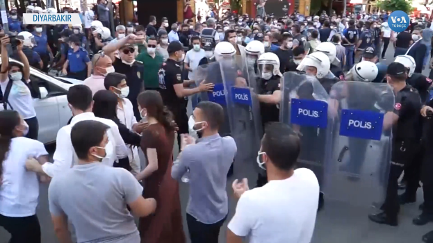 Eine Gruppe von Menschen steht vor einer Reihe von Polizisten in Helmen und Schilden, mit einem Auto links, einer Person mit einer Kamera rechts und Gebäuden, Bäumen und Texttafeln im Hintergrund in Ankara, Türkei.
