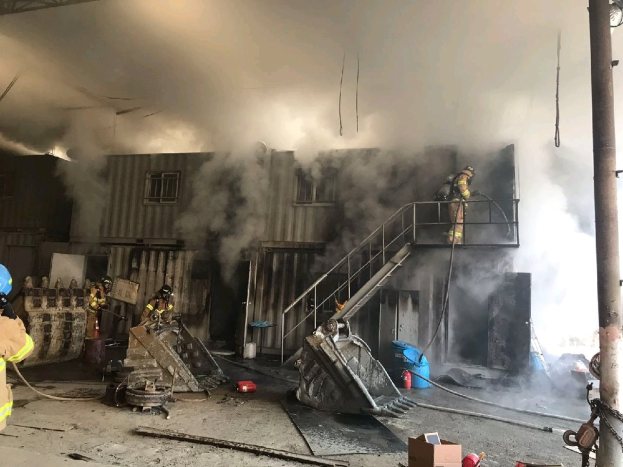 Feuerwehrleute in Helmen und Schutzausrüstung spritzen Wasser aus Schläuchen, um ein Lagerfeuer zu löschen, mit dicken Rauchwolken, die aus dem Gebäude aufsteigen und Trümmer auf dem Boden verstreut sind.