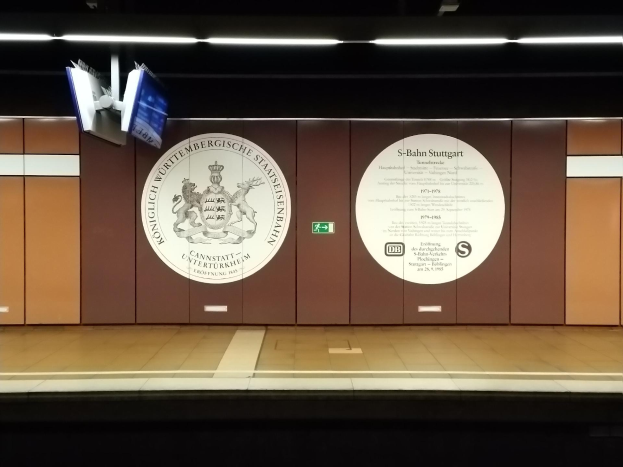 Ein Bahnsteig mit einem Schild, auf dem 'S-Bahn Stuttgart' steht und ein Logo an der Wand, einige Lampen an der Decke und einige Gegenstände auf der linken Seite.