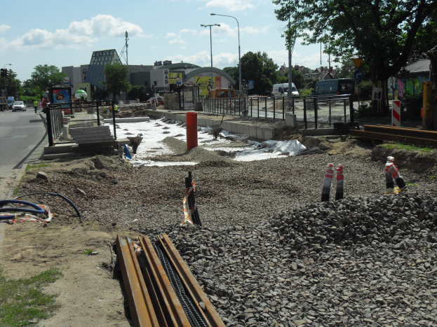 Baustelle mit verschiedenen Materialien und Geräten entlang einer Straße, Fahrzeuge und Gebäude im Hintergrund unter einem bewölktem Himmel.