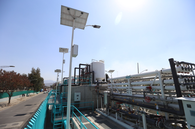 Großes industrielles Gebäude mit ausgedehnten Rohrleitungen und Solarmodulen auf dem Dach, umgeben von Sicherheitsgeländern, Strommasten, Straßenlaternen, Schildern, Bäumen und vorbeifahrenden Fahrzeugen, vor einem Hügelpanorama unter einem klaren blauen Himmel, Teil der neuen Wasseraufbereitungsanlage des California Department of Environmental Protection.