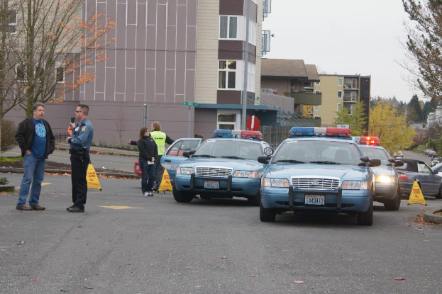 Autos auf einer Straße mit vier Menschen in der Nähe, Gebäude mit Fenstern im Hintergrund, Bäume und Warndreiecke.