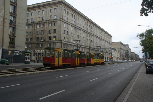 Eine Stadtansicht mit Gebäuden, Fahrzeugen, einer Bahn, Straßenlaternen, Pfählen, Kabeln, einer Straße, Bäumen, Fenstern, Geschäften und einer bewölkten Himmel.