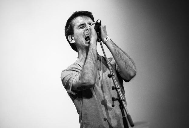 Ein Mann singt in ein Mikrofon auf einer Bühne, trägt ein schwarzes und weißes Outfit und hat ein Mikrofonständer vor sich.