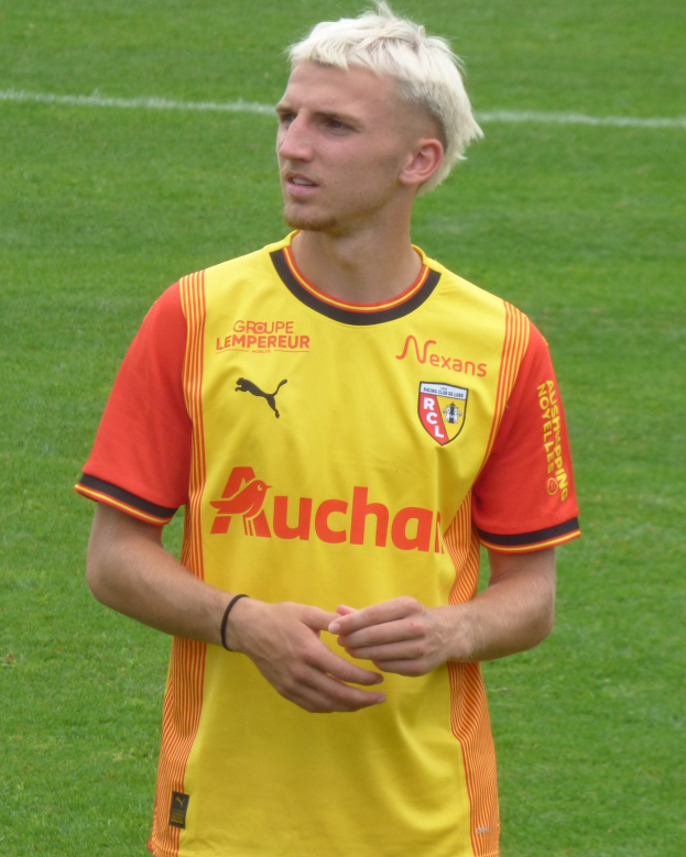 Ein Mann in einem gelben und orangefarbenen T-Shirt steht selbstbewusst auf einem Fußballfeld und sieht aus wie ein Flügelspieler mit entschlossener Miene.