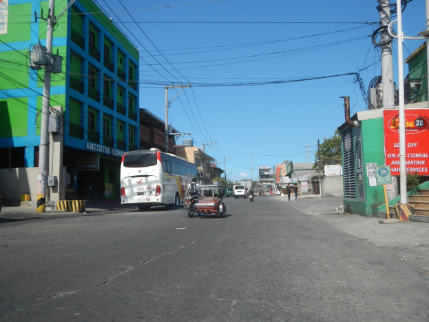 Eine Stadtstraße mit einem Bus, der daran vorbeifährt, flankiert von hohen Gebäuden, Strommasten mit Drähten, einigen Fußgängern auf dem Gehweg und Bäumen unter einem klaren blauen Himmel im Hintergrund.