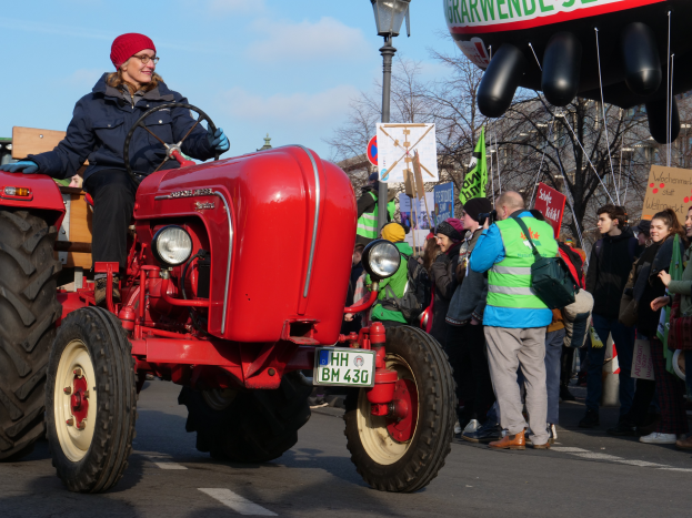 Eine Frau fährt einen roten Traktor die Straße entlang, umgeben von einer Menge mit Schildern und Transparenten, mit Laternen, Bäumen, Gebäuden und einem klaren blauen Himmel im Hintergrund.