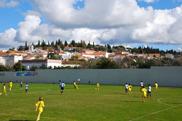 Eine Gruppe von Menschen, die ein Spiel auf einem Feld mit Plakaten an einer nahen Wand, Bäumen, Pfählen, Häusern und Wolken im Hintergrund spielen.