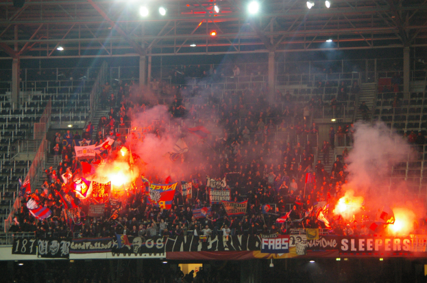 Eine große Menge in einem Stadion hält Fahnen und Banner, mit Leuchtfackeln, die Rauch erzeugen, unter einer Decke mit Deckenleuchten und Metallrahmen.
