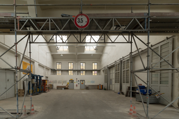 Lagerinneres mit Gerüsten, Verkehrskegeln, Metallstangen, einem Schild, Maschinen, einer Tür, Fenstern, einer Wand, Deckenlampen und Dach.