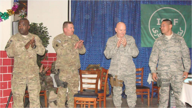 Vier Personen stehen, drei klatschen, eine sitzt auf einem Stuhl neben einer Pflanze, mit einem Banner im Hintergrund, alle tragen militärische Kleidung, Stühle und ein Tisch sind sichtbar.