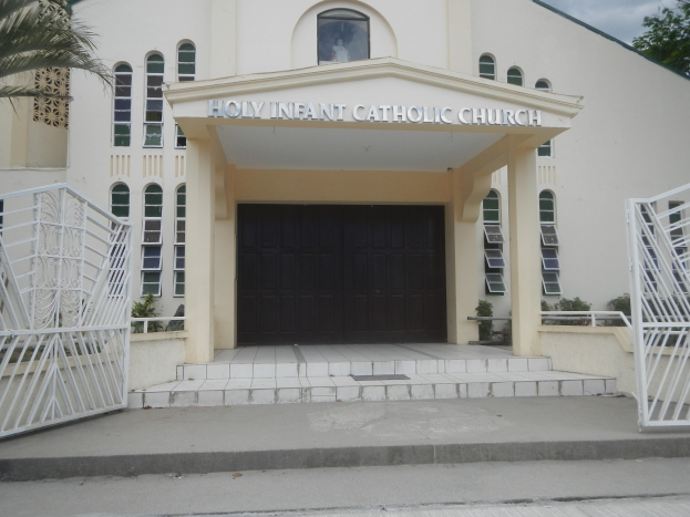 Außenansicht der Heiligen-Kind-Katholischen Kirche mit Treppe, Geländer, umgebender Vegetation und einem bewölkten Himmel.