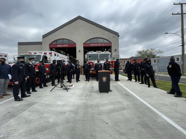 Gruppe von Menschen in Kappen in der Nähe eines Feuerwehrautos auf einem Podium während einer Feuerwachen-Einweihungszeremonie, mit Gebäuden, Bäumen, Strommasten und Gras im Hintergrund unter einem bewölkten Himmel.