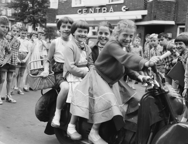Schwarzes und weißes Bild von Kindern, die auf dem Rücksitz eines Motorrollers fahren, mit zwei sitzenden und einem mit einem Korb haltenden, während andere auf der Straße im Hintergrund mit Gebäuden, Bäumen und einem Pfahl stehen.