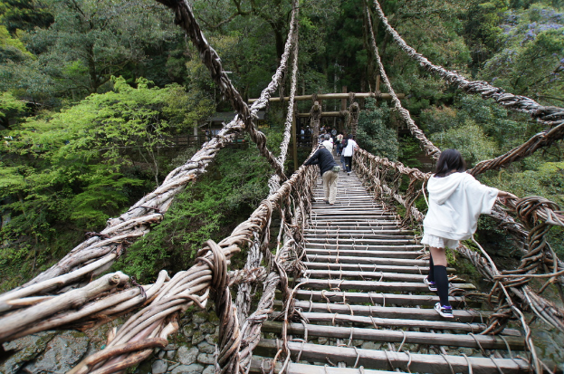 Eine Brücke mit wenigen Fußgängern, umgeben von Bäumen, Pflanzen, Steinen, einer Hütte und einem Zaun.