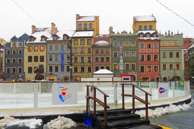 Eislaufbahn in einem Stadtplatz umgeben von Gebäuden, Treppen, Geländern, Informationsschildern, Schnee, Laternen, Bäumen und Himmel.