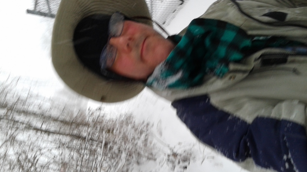Eine Person in Jacke und Hut steht vor einem verschneiten Hintergrund mit Bäumen im linken unteren Eck.