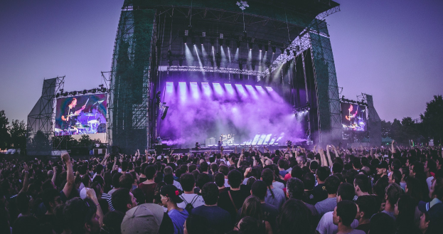 Eine große Menschenmenge auf einem Musikfestival schaut auf einer hell beleuchteten Bühne mit Instrumenten und Bildschirmen, die vor einem Gebäude, Bäumen und einem klaren Himmel aufgebaut sind.