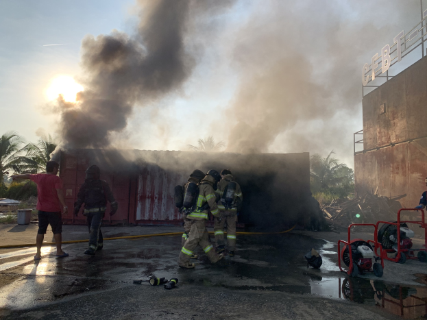 Feuerwehrleute in Helmen stehen vor einem brennenden Gebäude mit dicken Rauch, arbeiten daran, das Feuer zu löschen; ein benanntes Gebäude ist rechts zu sehen, und Bäume und andere Gegenstände links.