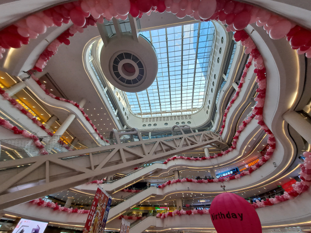 Ein Einkaufszentrum-Innenraum dekoriert für den Valentinstag mit pinken und weißen Luftballons, Rolltreppen, Geländern, Texttafeln, Lichtern und einer Glasdecke.