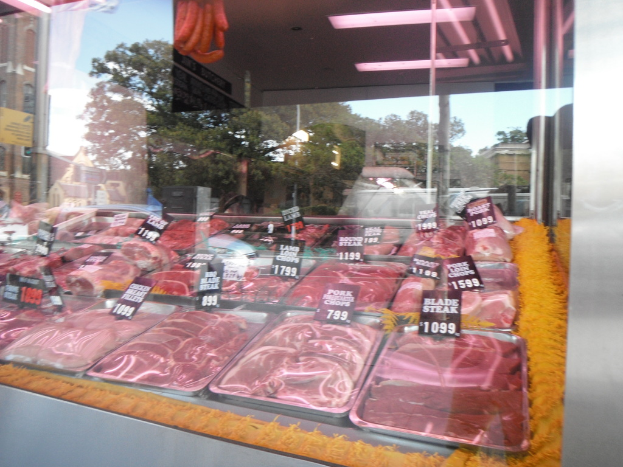 Eine Glasvitrine in einem Fleischgeschäft, die Tabletts mit Fleisch und Preisschildern zeigt, mit Gebäuden und Bäumen im Fenster und der Decke des Raums oben.