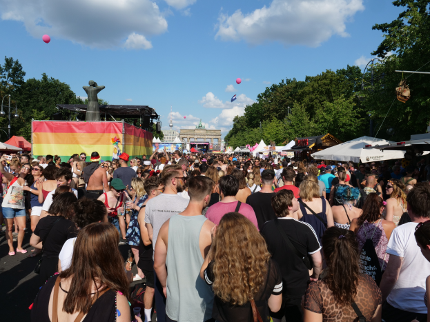 Eine große Menschenmenge, die eine Straße mit Zelten, Bäumen, Pfählen, Laternen und einem Gebäude im Hintergrund entlanggeht, bei einem Fest in Berlin.