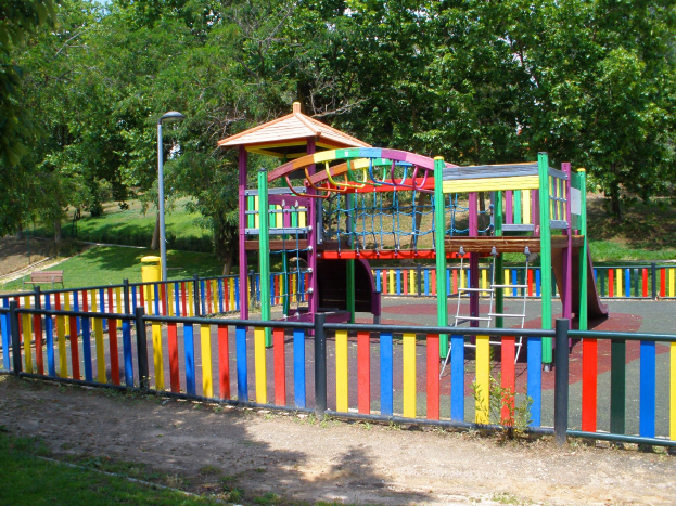 Ein Spielplatz mit einer bunten Spielstruktur in der Mitte, umgeben von einem Zaun, Pfählen, Laternen, Bänken, Gras, Pflanzen und Bäumen, unter einem klaren blauen Himmel.