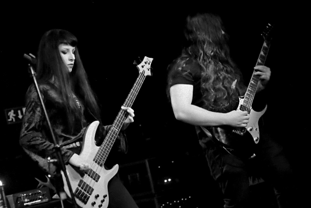 Zwei Frauen in schwarzen Kleidern, die auf der Bühne Gitarren spielen, mit einem Mikrofon davor und verschiedenen Musikinstrumenten drumherum.
