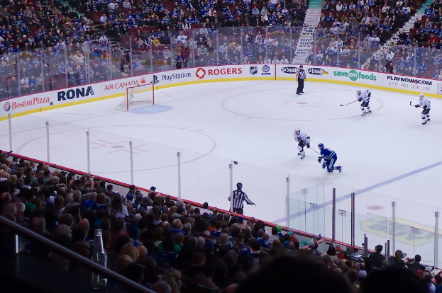 Vier Männer beim Eishockeyspielen auf einem Eis, mit zwei in der Nähe des Tors und Zauns stehend, Zuschauern auf Stühlen im Vorder- und Hintergrund und einer Treppe im Hintergrund zu sehen.