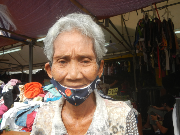 Alte Frau mit Gesichtsmaske in einem Markt umgeben von Körben, Lichtern, Ventilatoren und anderen Gegenständen mit einer Person im Hintergrund sitzend.