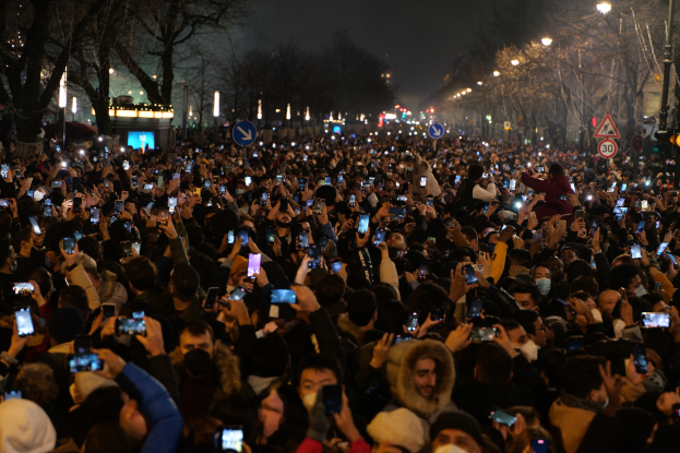 Eine große Menschenmenge steht nachts auf einer Straße, beleuchtet von Straßenlaternen, umgeben von Bäumen und Schildern, viele halten Mobiltelefone hoch.