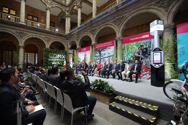 Eine Gruppe von Menschen sitzt in Stühlen und schaut auf einen Mann auf einer Bühne, der ein Mikrofon hält, mit Fahrrädern, die rechts geparkt sind, und verschiedenen Elementen wie Plakaten, Pflanzen, Säulen, Geländern, Lampen, Fenstern und einer Wand im Hintergrund, wahrscheinlich bei einer Pressekonferenz für den CDMX-Nachhaltigkeitsenergiegipfel.