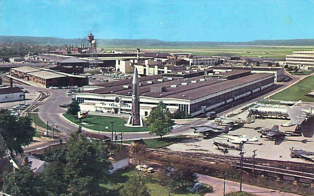 Altes Schwarz-Weiß-Foto eines Flughafens mit geparkten Flugzeugen, Gebäuden, Bäumen, Straßen, Fahrzeugen, Strommasten, Gras, Bahnschienen, Türmen, fernen Hügeln und einem klaren Himmel, mit Text unten.