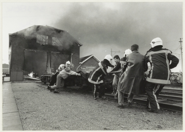 Firefighters in helmets stand near a train as a house burns in the background, smoke rising into the sky.