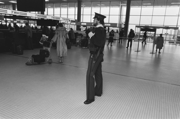 Ein Polizeibeamter in Uniform steht in einem Flughafen-Terminal, hält eine Waffe und ist von Menschen und Gepäckwagen umgeben, mit Masten, Texttafeln, Deckenlampen und Glaswänden im Hintergrund.