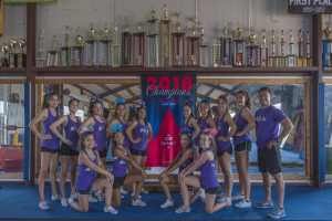 Eine Gruppe von Cheerleadern in lila T-Shirts und schwarzen Shorts posiert vor einem Vitrinenschrank mit einer "2018 Summit Cheerleading Champions"-Banner im Hintergrund.