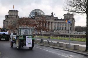 Eine Gruppe von Traktoren fährt die Straße vor dem Reichstaggebäude in Berlin, Deutschland, entlang. Das Gebäude ist mit Fenstern, Säulen und Flaggen geschmückt und von Bäumen, Geländern, Pfosten, Brettern und anderen Gegenständen umgeben.