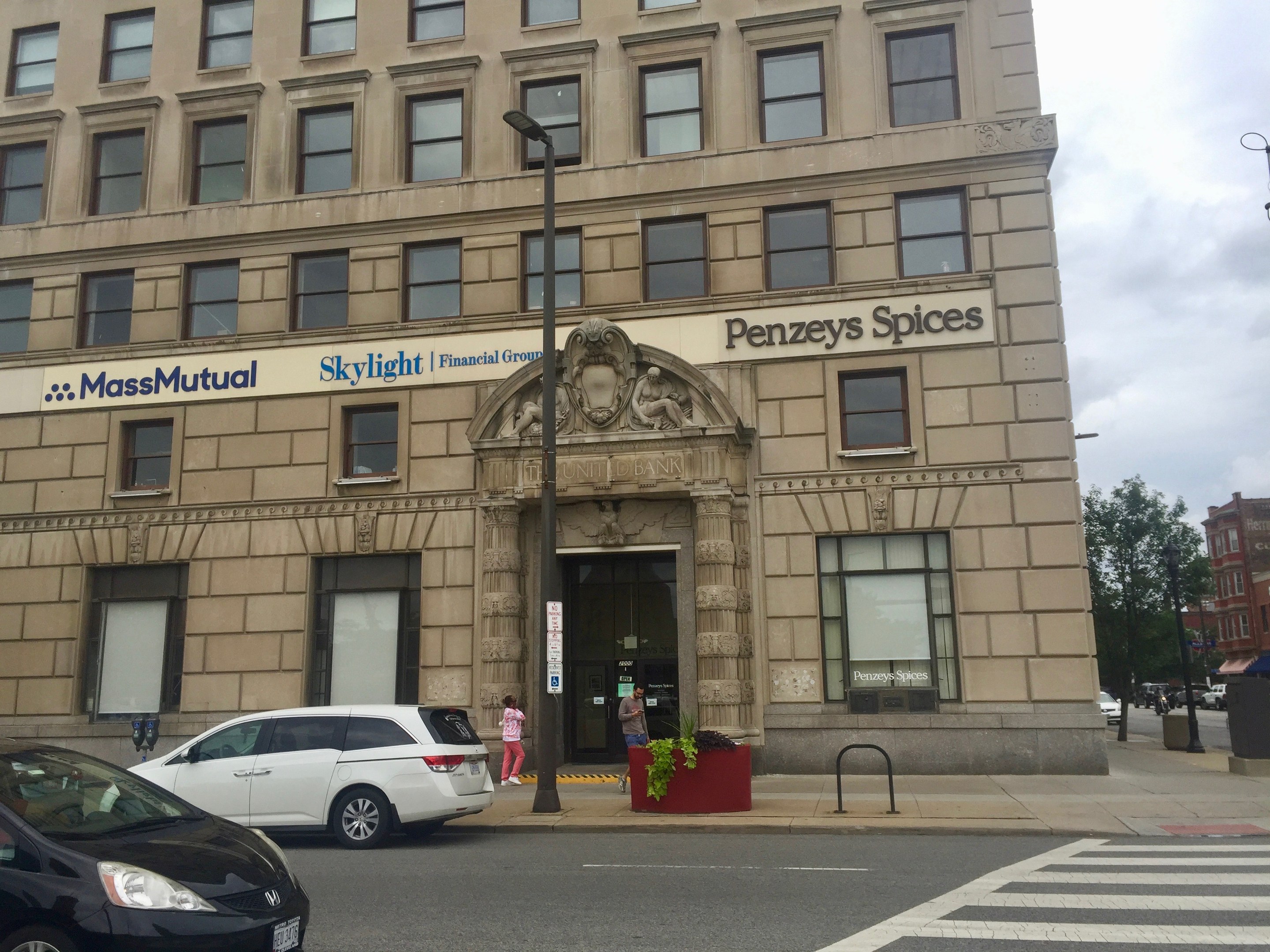 Großes Gebäude mit parkenden Autos davor, Straßenlaternen, fahrende Autos, Fußgänger auf dem Gehweg, Grünfläche und bewölkter Himmel, identifiziert als Hauptsitz der Massachusetts Mutual Financial Group.