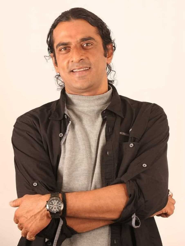 Ein Mann in einem schwarzen und aschfarbenen Kleid mit verschr├Ąnkten Armen, der eine Uhr tr├Ągt und vor einem wei├čen Hintergrund steht, in Gedanken versunken.