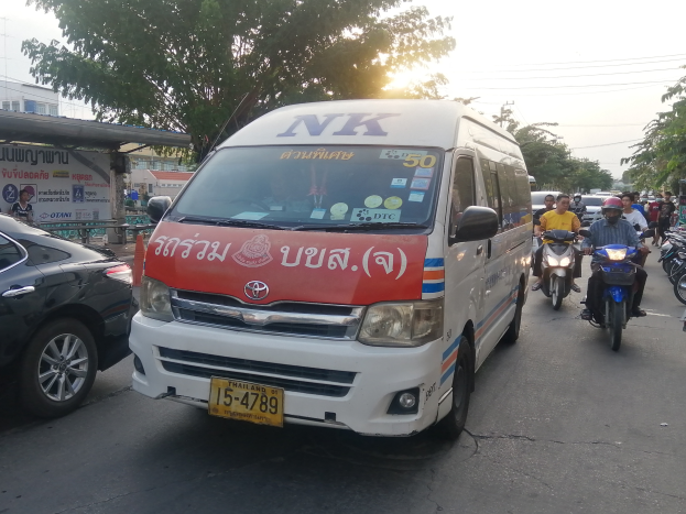 Ein weißer Lieferwagen fährt eine Straße mit parkenden Autos, Motorrädern und Menschen mit Helmen entlang, mit Bäumen, Gebäuden, Texttafeln, Polen, Drähten und einem klaren blauen Himmel im Hintergrund.