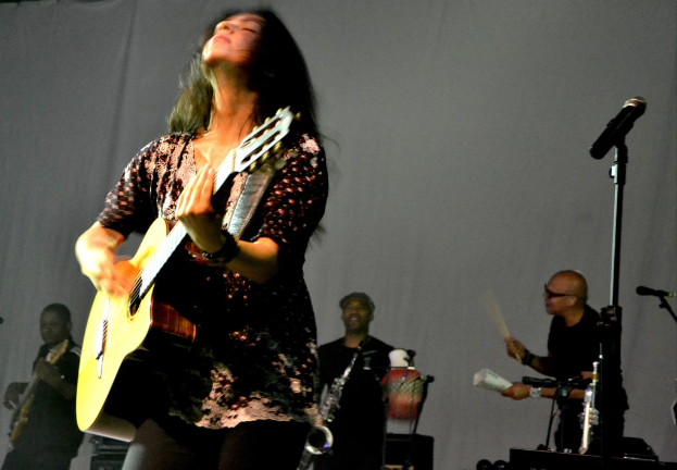 Eine Frau, die auf der Bühne eine Gitarre spielt, mit Musikern und verschiedenen Instrumenten im Hintergrund und einem Mikrofon rechts daneben.